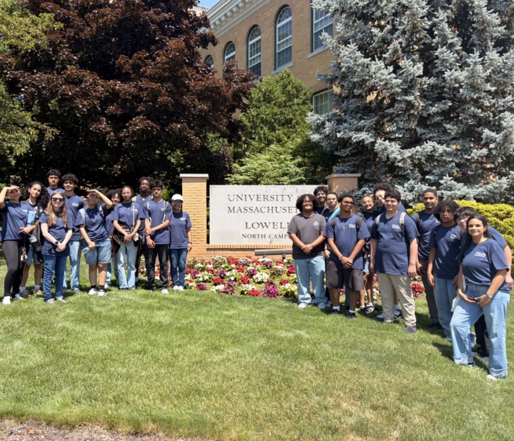 students pose smiling at the camera in front of the UMass Lowell sign