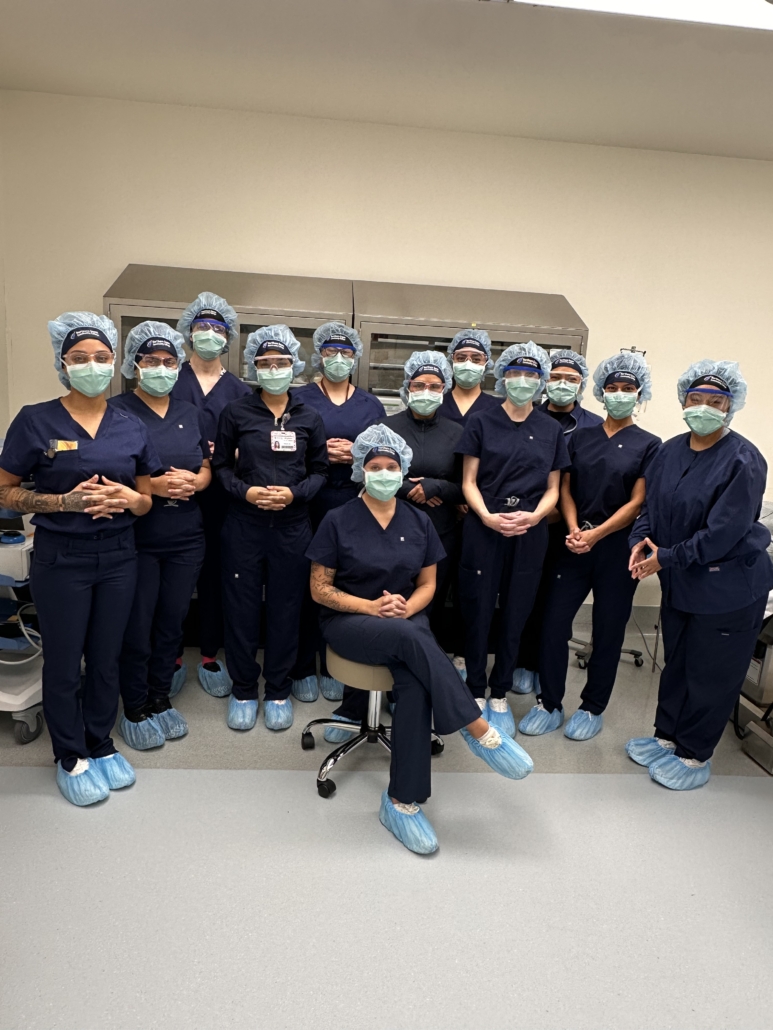 group shot of surgical tech students wearing scrubs, face masks and hair covers. They stand posed in OR looking at the camera