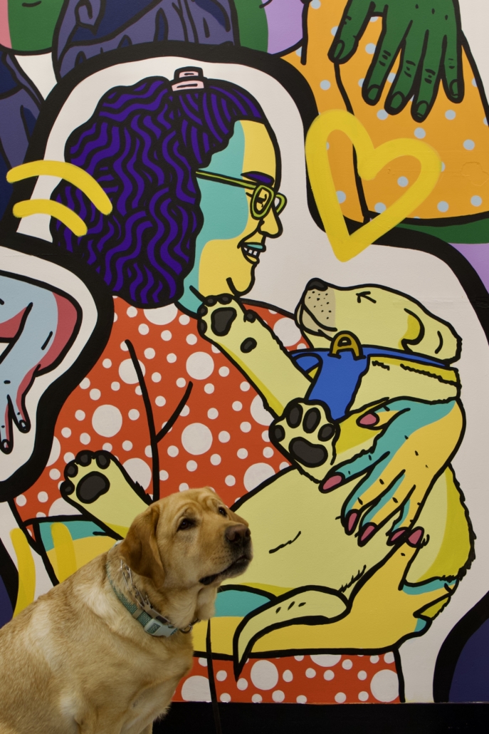 Winnie the yellow lab poses in front of a drawing of her as a puppy