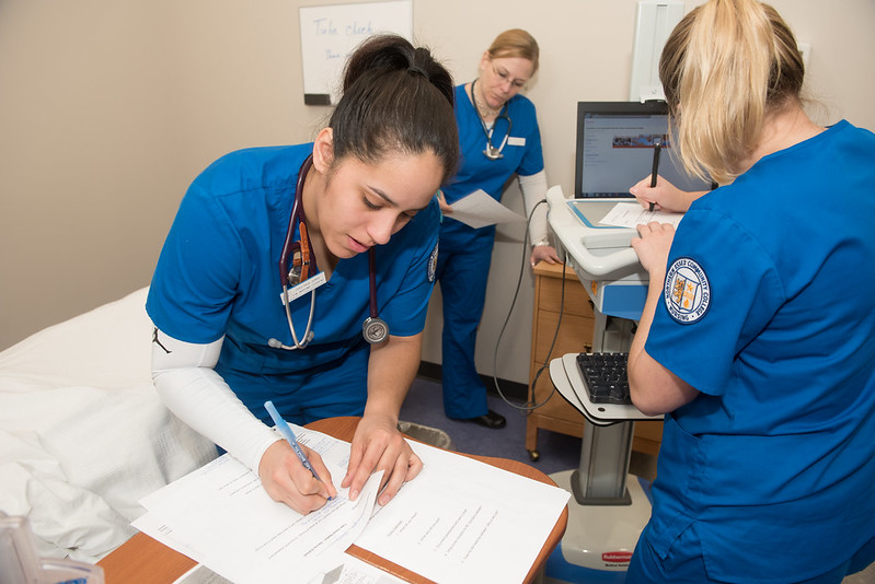 three nursing students fill out paperwork, all three are wearing blue scrubs