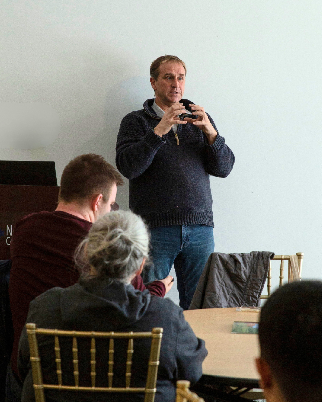 dale rogers, wearing a sweater and jeans, stands to the left of a lecturn and gestures with his hands to the seated audience
