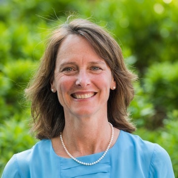 marjorie, in light blue top, standing in front of greenery, smiles at camera