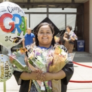 student in cap and gown smiles at the camera, her arms full of flowers and balloons
