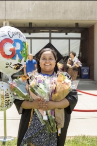 student in cap and gown smiles at the camera, her arms full of flowers and balloons