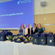 members of the HR staff stand behind a table full of gift bags. Behind is a screen with the words "Service Recognition Lunch"