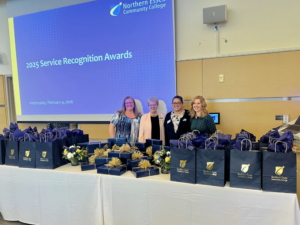 members of the HR staff stand behind a table full of gift bags. Behind is a screen with the words "Service Recognition Lunch"