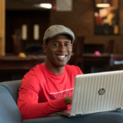 student in a red shirt sits smiling from behind a laptop