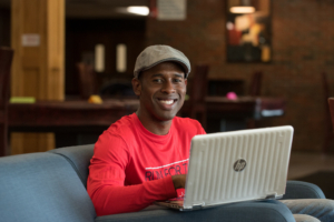 student in a red shirt sits smiling from behind a laptop