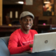 student in a red shirt sits smiling from behind a laptop