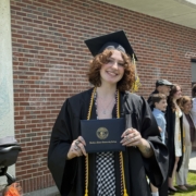 mckenzie stands wearing cap and gown at commencement, holds her diploma