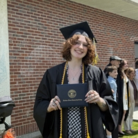 mckenzie stands wearing cap and gown at commencement, holds her diploma