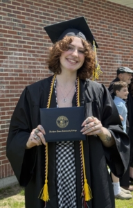 mckenzie stands wearing cap and gown at commencement, holds her diploma