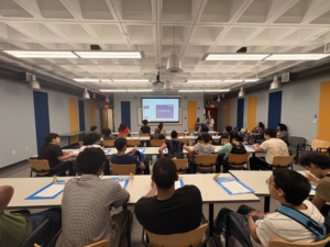 a group of students sit in a classroom, looking ahead at a powerpoint presentation