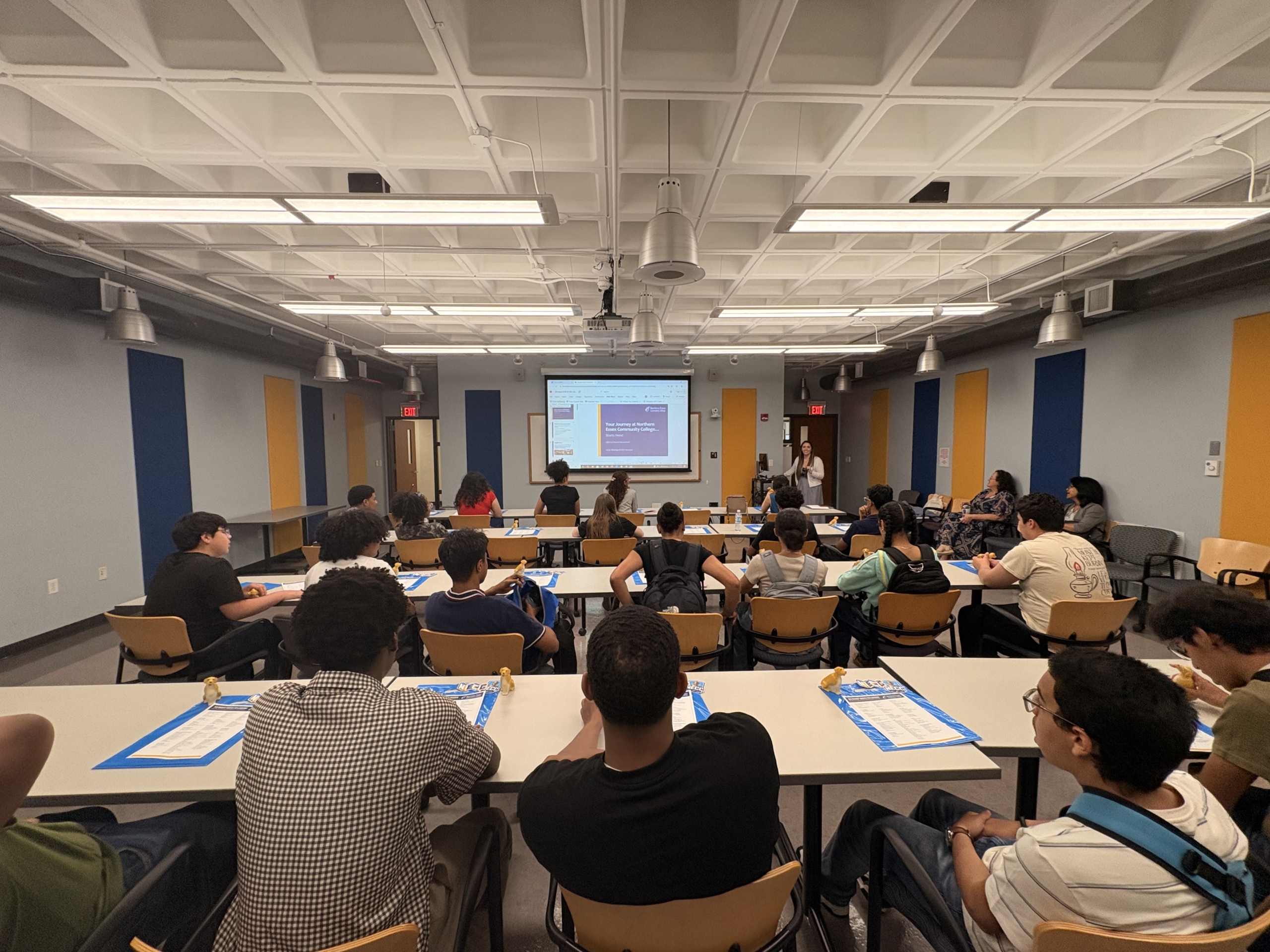 a group of students sit in a classroom, looking ahead at a powerpoint presentation