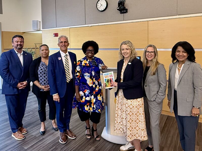 michaela, wearing a colorfully printed dress, stands with her book. She is posing with NECC President Lane Glenn and NECC Vice Presidents