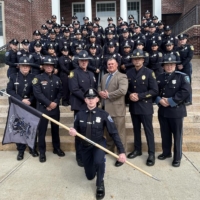 new officers pose in their dress blues on the steps outside of a brick building. One kneels in front holding the MPTC flag