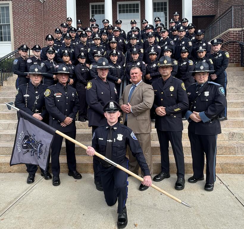 new officers pose in their dress blues on the steps outside of a brick building. One kneels in front holding the MPTC flag