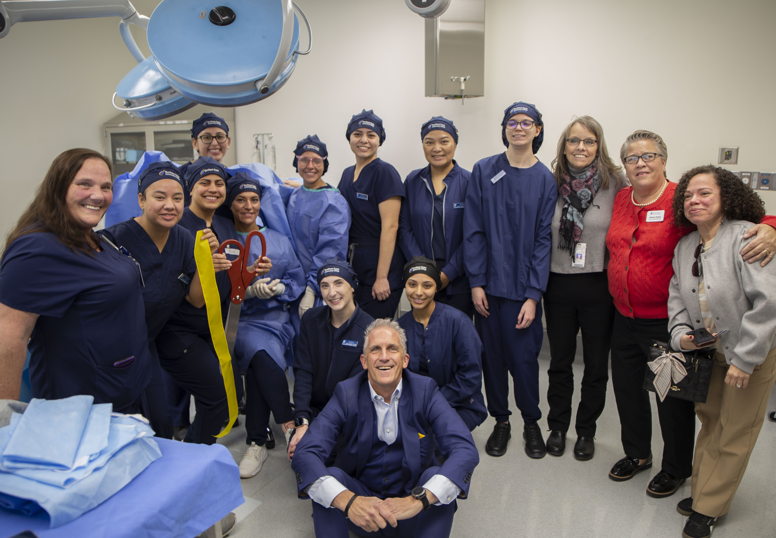 a group of students in scrubs stand posing in the OR, president Lane Glenn sits on the floor in front of them