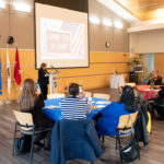 wide shot of room with tables and woman speaking at a podium