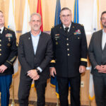 Four men pose in front of flags smiling