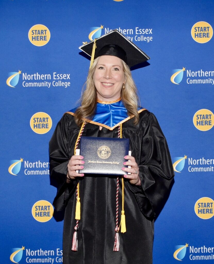 kristie poses in front of backdrop with NECC logo, in her graduation cap and gown, holding her diploma
