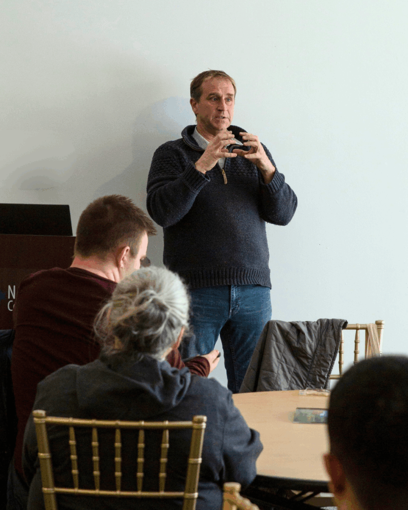 dale rogers, wearing a sweater and jeans, stands to the left of a lecturn and gestures with his hands to the seated audience 
