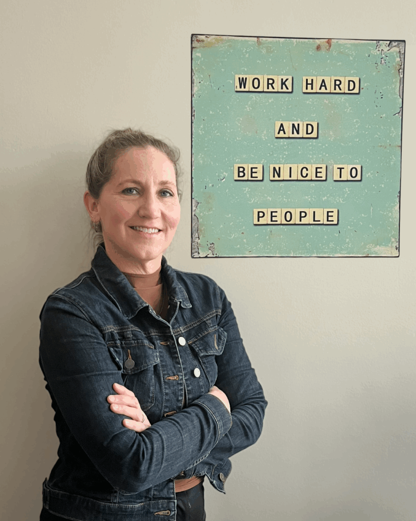 Lori Capr stands against office wall with poster behind reading "work hard and be nice to people"