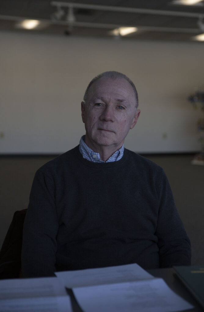 photo of NECC instructor Kevin Keleher. he sits in a chair in a dimly lit room with a white background. the photo is taken from the waist up