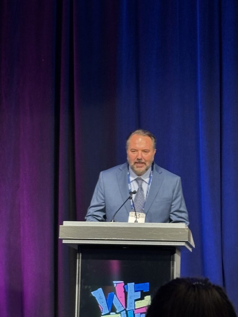ron, in a suit and tie, stands at a podium with purple draping behind him