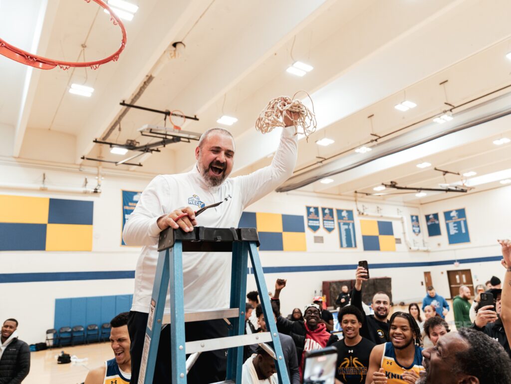 head coach darren straton stands on ladder, cutting part of the net from the basket