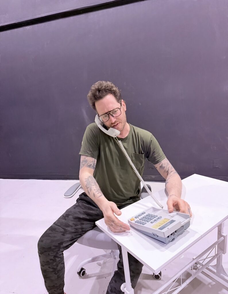 Actor Michael Zuccola speaks on a corded phone, seated at a small white desk