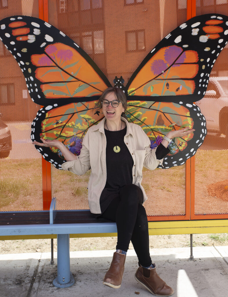 Professor Jenn Houle sits in the bus shelter on the bench in front of her butterfly mural
