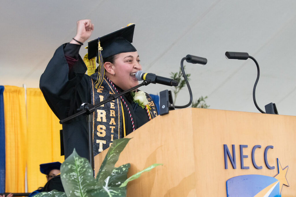 maria stands behind a podium and microphone, her hand raised in excitement
