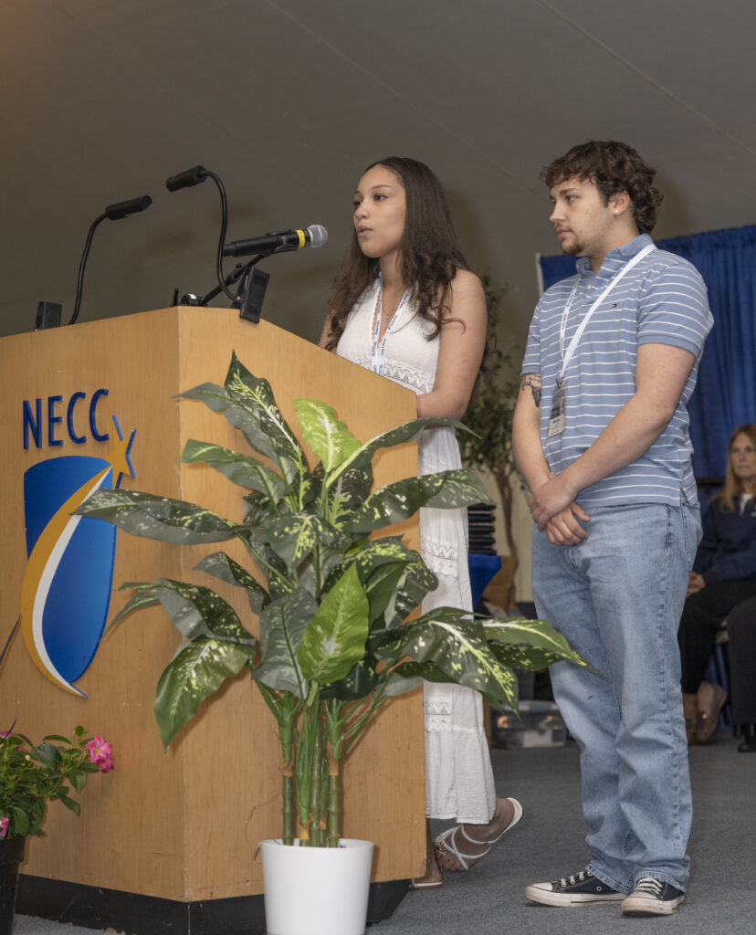Two students stand behind podium with microphone and NECC Logo
