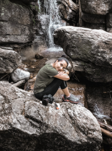 Alejandra sits on a rock in front of a waterfall with her camera at her side.