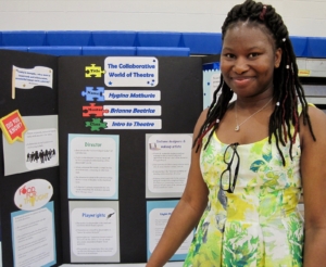 Young Black woman in floral dress stands in front of her honors project titled The Collaborative World of Theater.