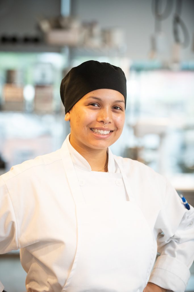 image of culinary student Mercedes Rodriguez in chef's coat and apron, standing hands on hip smiling at the camera