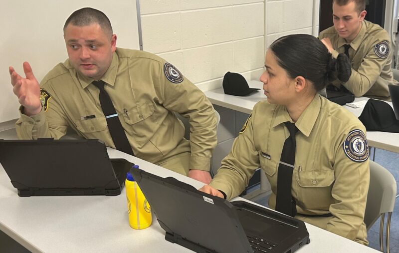 Three participants in the program, a male presenting, a female presenting, and a male presenting in the background. Sitting at desks, in uniform.